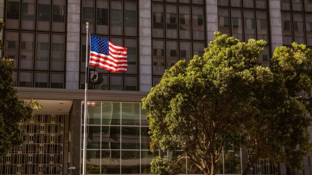 A vibrant American flag waves in front of a modern building facade amidst trees.
