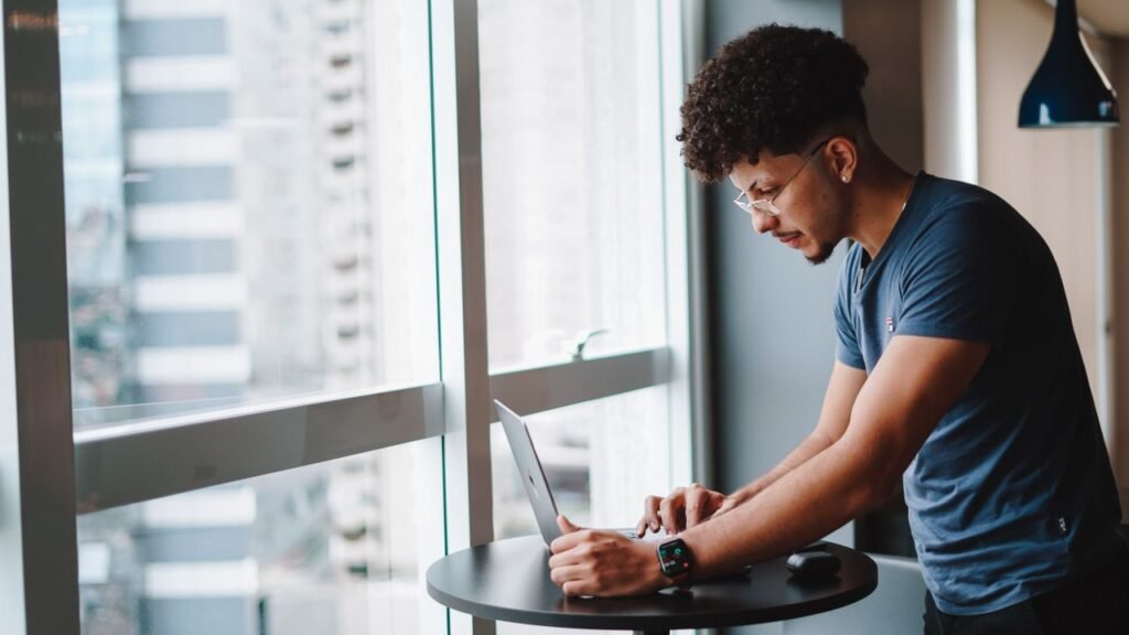 5 Careers Gen Z in America Actually Wants Right Now Young man using laptop standing by window in modern office setting.