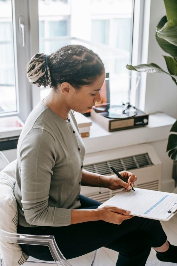 Side view of concentrated ethnic female psychologist sitting with crossed legs and taking notes on clipboard during work in office