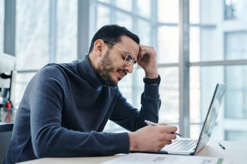 7 Signs You're Underpaid (And What to Do About It) Man looking stressed at laptop in a modern office with large windows.