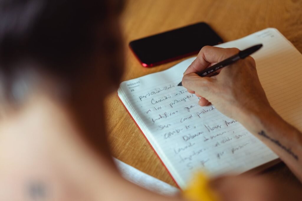 A person writes in a notebook at a wooden desk with a phone nearby.