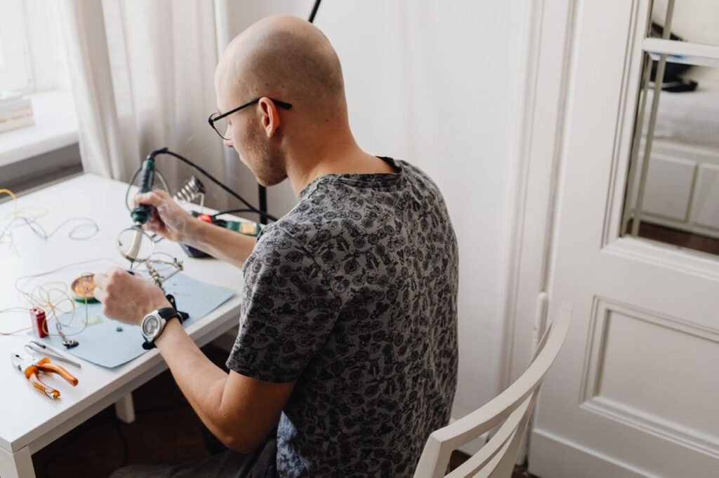 Bald man working at a desk with electronics, using a soldering iron, in a bright indoor setting.