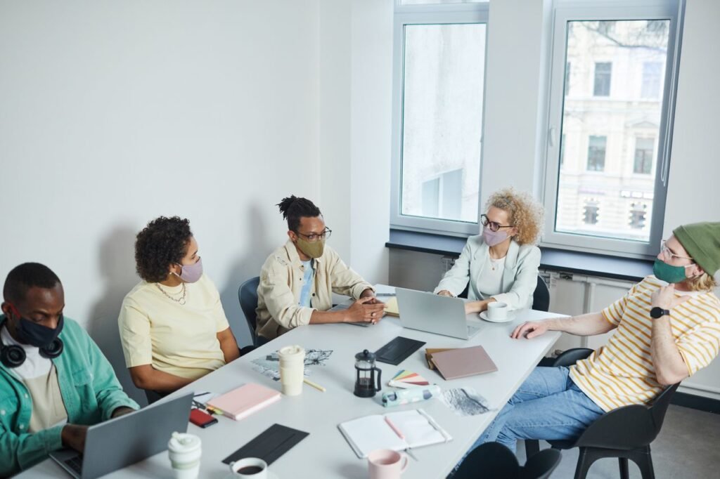 Remote, Hybrid, or In Office: Which Work Style Actually Fits You Office meeting with diverse colleagues wearing face masks, discussing projects in a modern workspace.