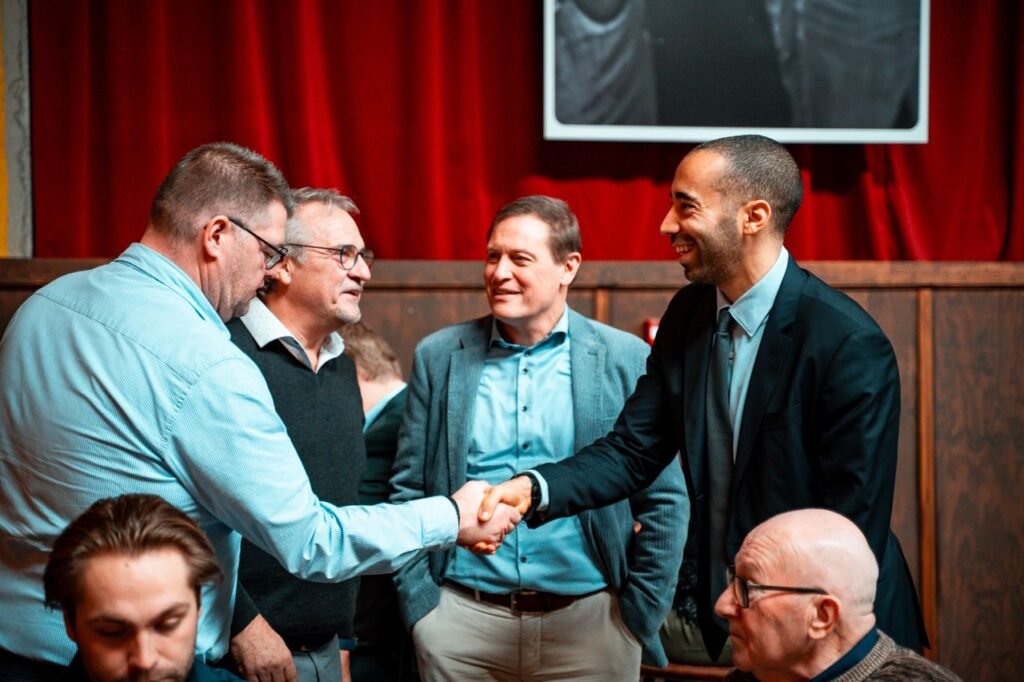 Five men in formal attire shaking hands and conversing at an indoor business meeting.